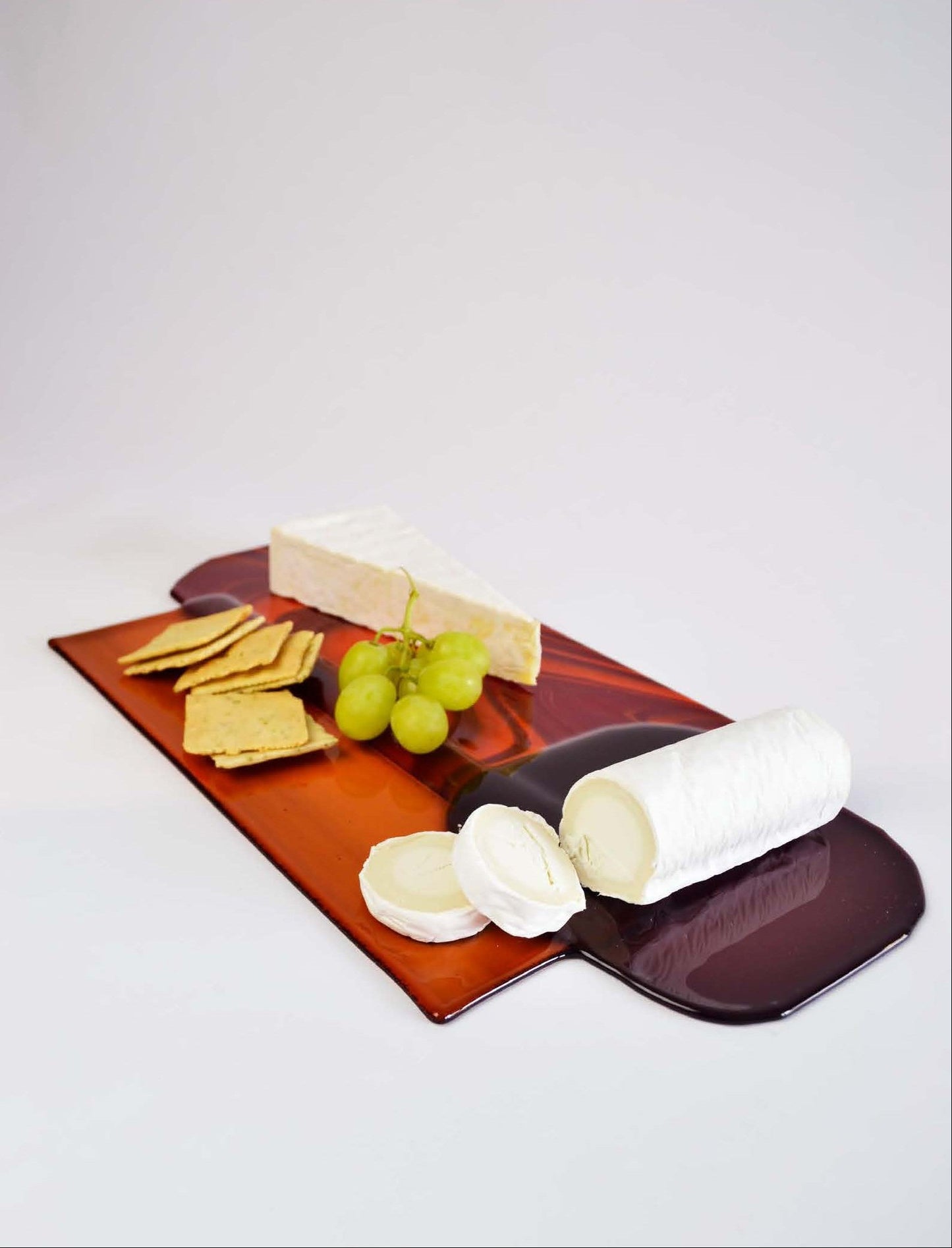 Artisan and handmade colored glass display plate. Used as cheese boards styled with cheese, crackers, grapes, and nuts on a light gray background.