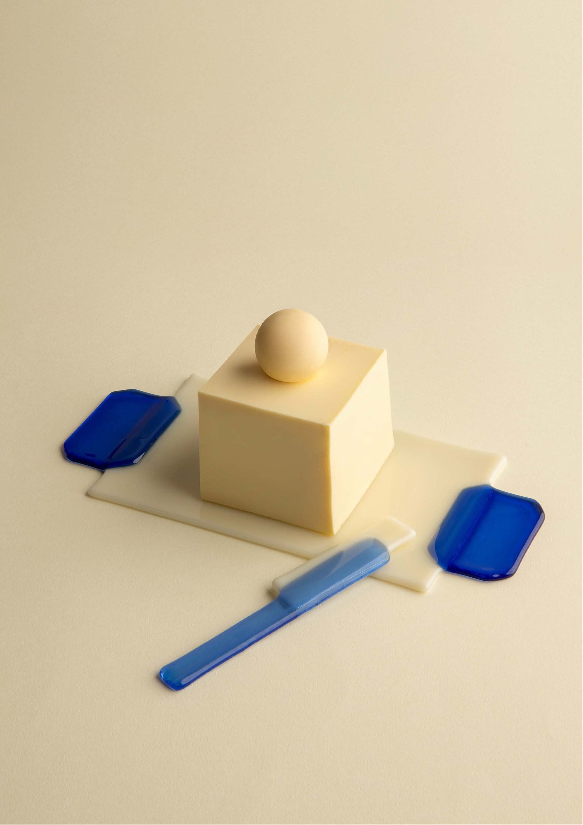A minimalist product photo showing a cream-colored butterplate and a matching butter knife with blue handles, arranged on a light beige surface next to a cream block and a small beige sphere. Perfect gift for your home