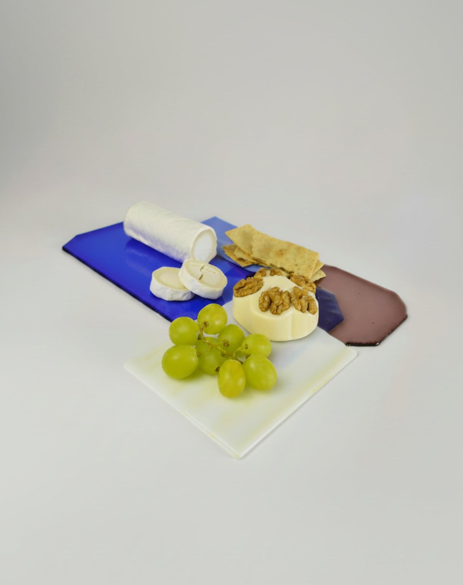 A styled food spread on a light gray studio background, featuring green grapes on a white square plate, a round cheese wheel topped with walnuts and crackers on a beige plate, a blue rectangular board with a white cylindrical container and sliced white food, and a brown oval board behind it.
Perfect gift for special ocassion and special house.